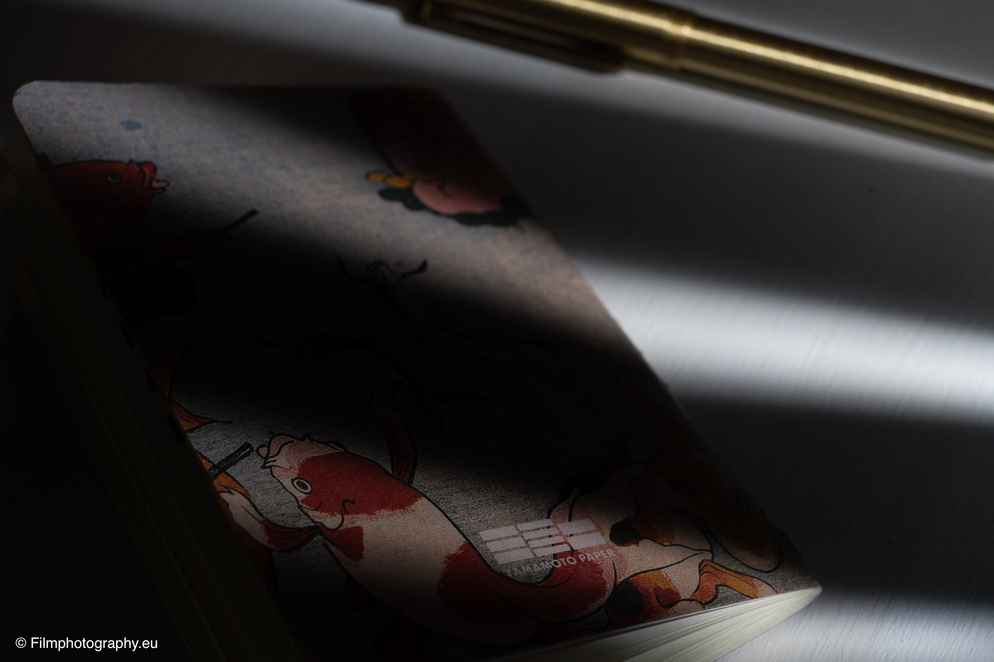 Yamamoto Robiki Notizbuch mit Koi-Fisch-Cover, Messingstift daneben, dramatische Lichtstrahlen und tiefe Schatten