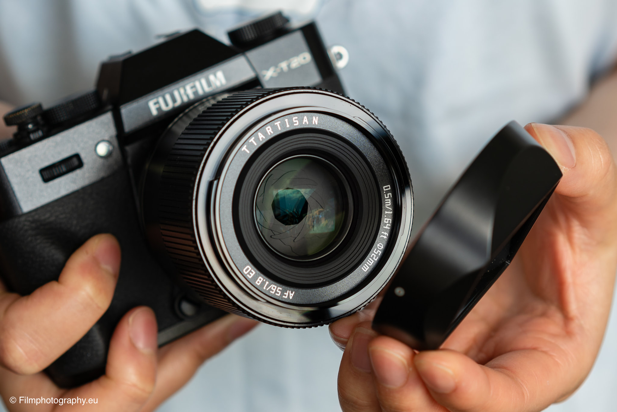 Nahaufnahme einer Fujifilm X-T20 mit TTArtisan 56mm f/1.8 Objektiv, Objektivkappe wird entfernt, weicher Stoffhintergrund.