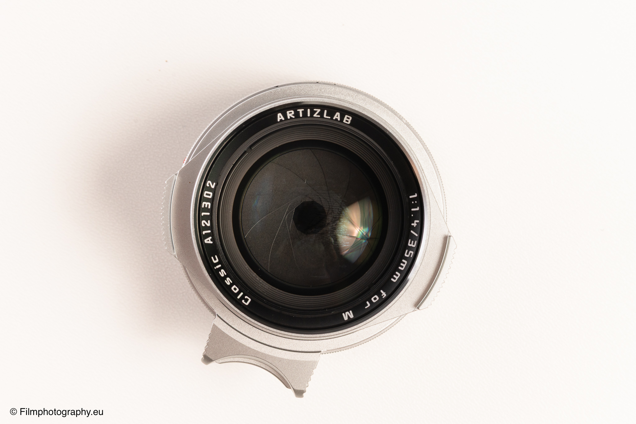 Frontansicht eines Artizlab Classic 35mm f/1.4 M-Mount Objektivs auf weißem Hintergrund, Deckel teilweise offen, Blende sichtbar.
