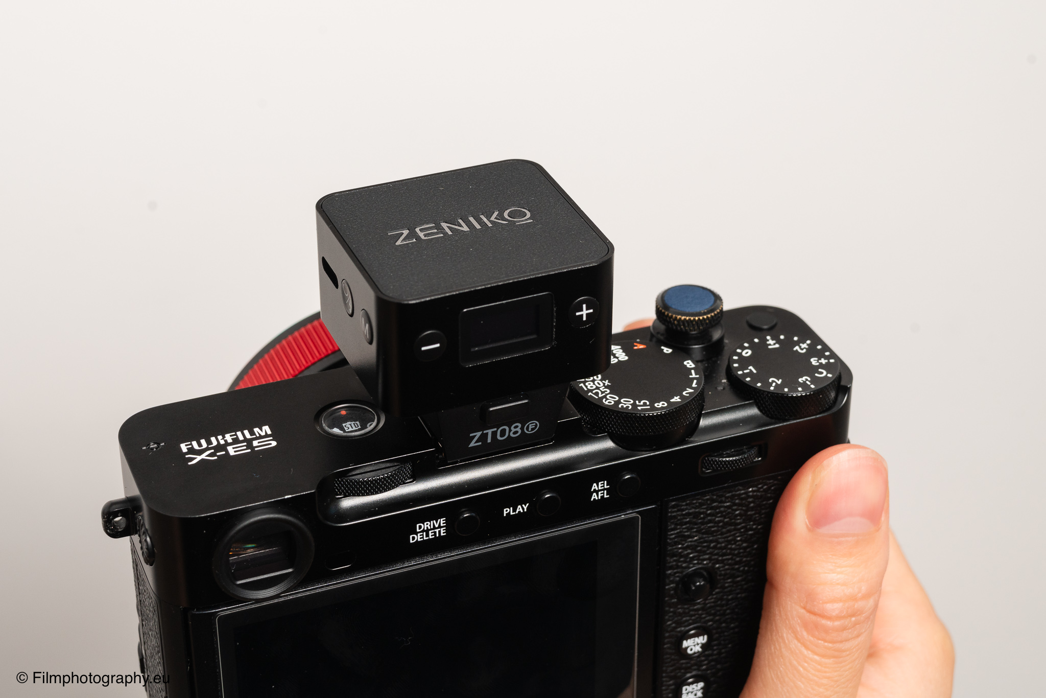 Fujifilm X-E5 mit Zeniko ZF08 TTL-Blitz oben montiert, in der Hand vor weißem Hintergrund mit klassischen Ziffernblättern.