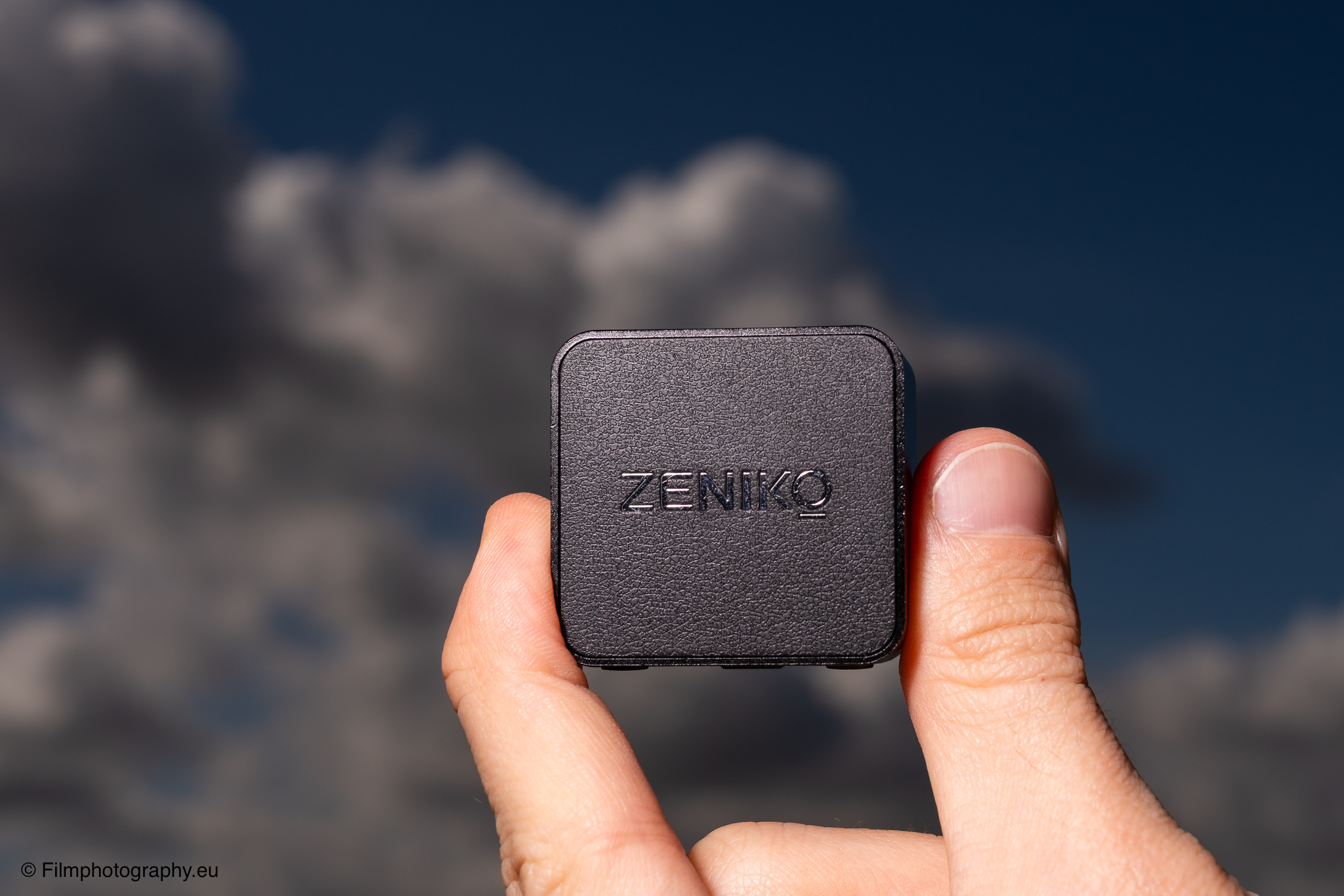 Hand hält einen Zeniko ZF08 TTL Blitzauslöser für Fujifilm-Kameras vor dramatischem bewölktem blauem Himmel