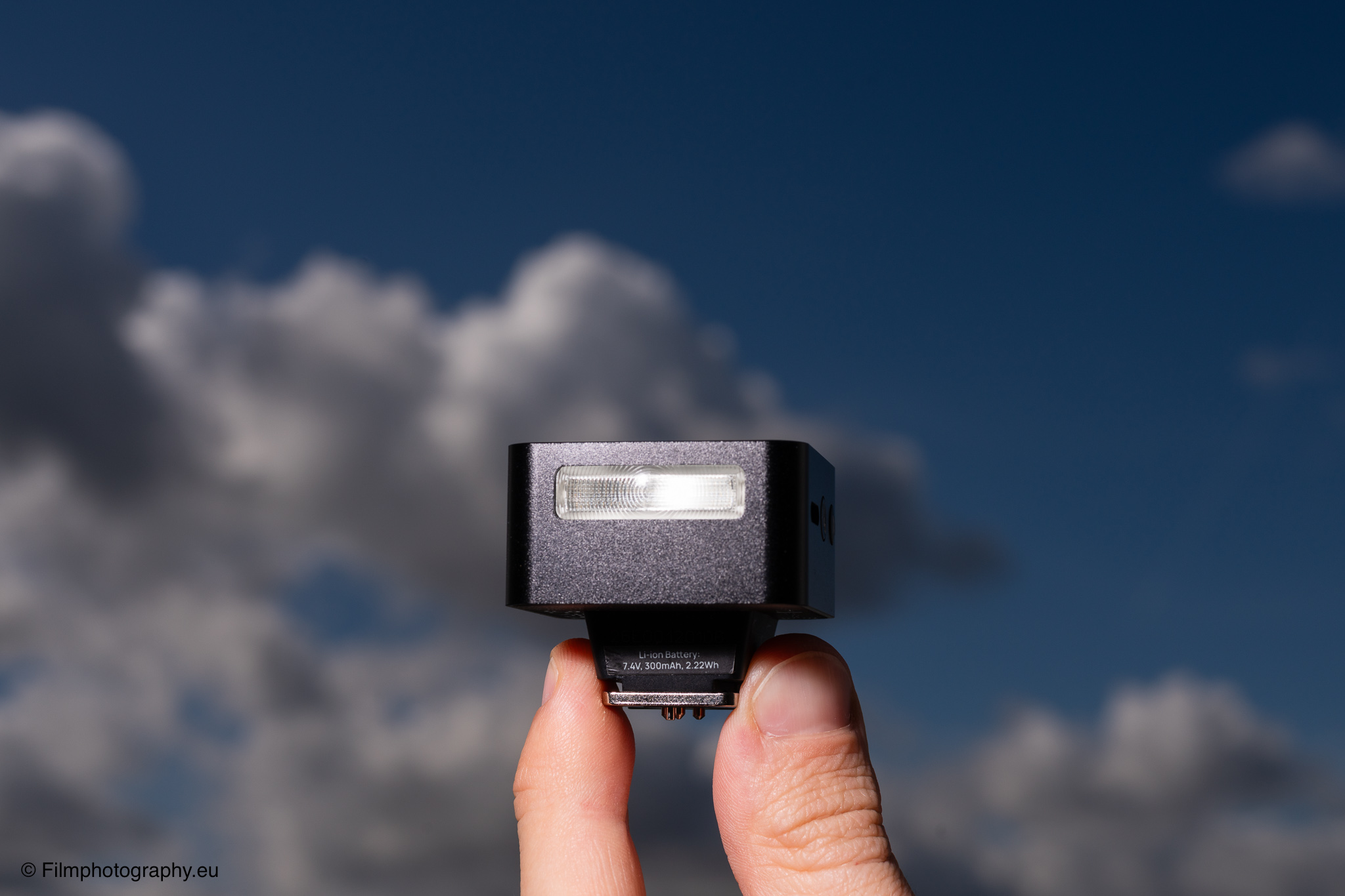 Hand hält eine Zeniko ZF08 TTL Blitzlicht für Fujifilm-Kameras vor einem hellblauen Himmel mit weißen Wolken