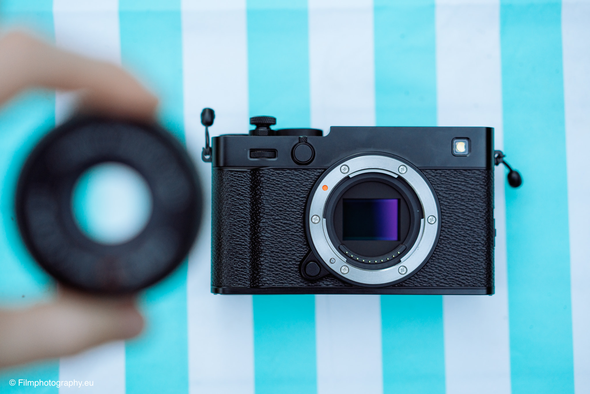 Schwarze Fujifilm X-E5 ohne Objektiv, Sensor sichtbar, auf türkis-weißem Grund mit unscharfem Deckel in Hand