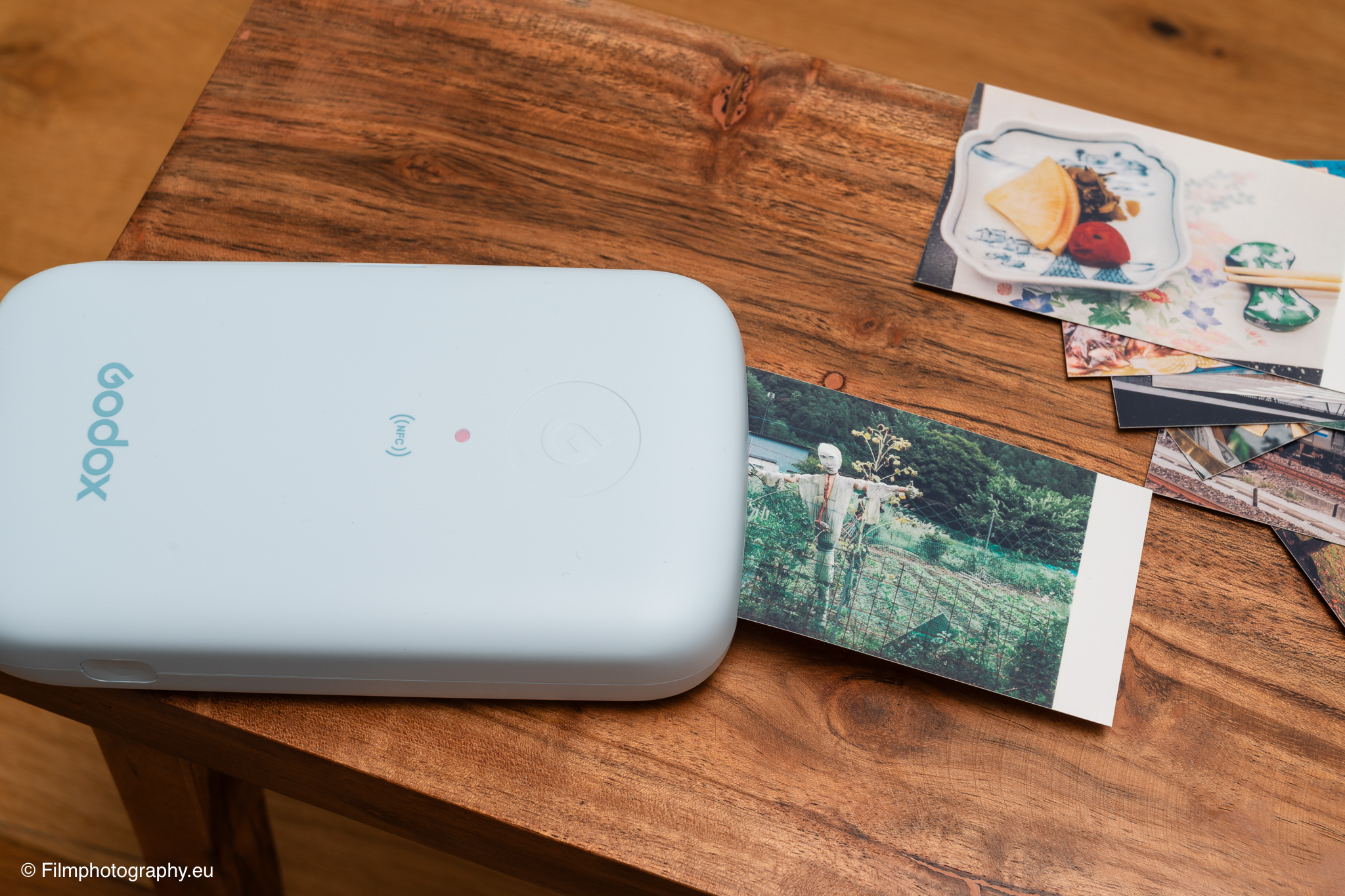 Godox P100 mobiler Fotodrucker auf einem Holztisch druckt ein Gartenfoto, gedruckte Schnappschüsse liegen verstreut daneben.