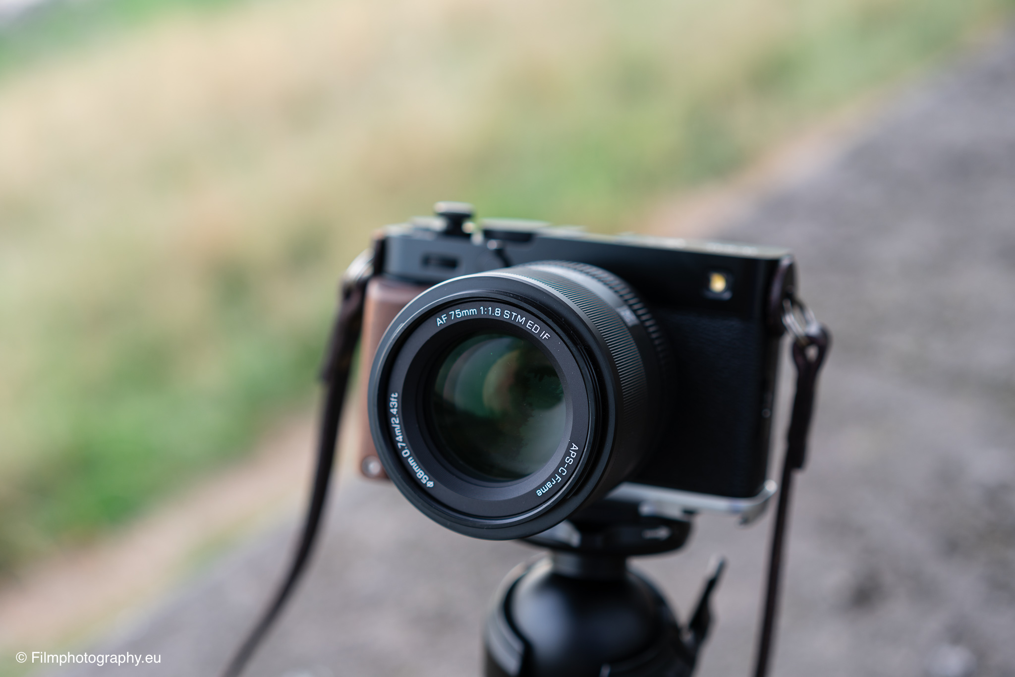 Fujifilm X-mount相机搭载Viltrox AF 75mm f/1.8镜头，置于三脚架上，背景为虚化的户外小径与绿色田野。